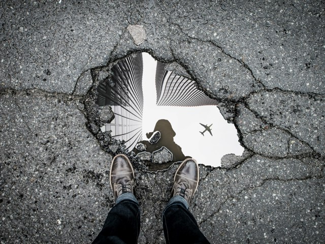 March 2026 Pothole with reflection of human, buildings and aeroplane (Unsplash/@marcojodoin/TQUERQGUZ8)